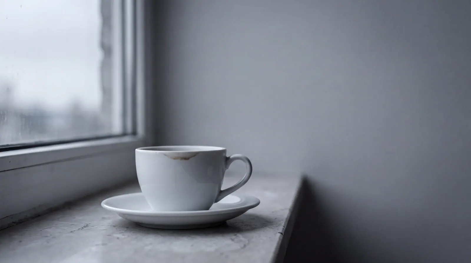 An empty white coffee cup and saucer on a pale stone windowsill in flat morning light