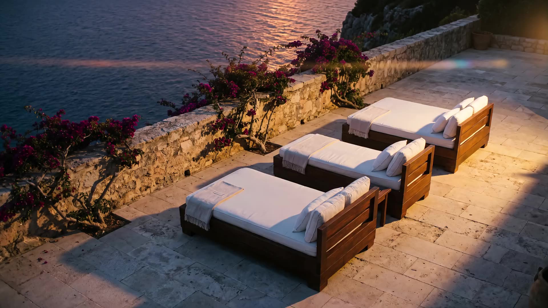 Linen sun loungers on a clifftop terrace above a calm sea, bougainvillea along the stone wall